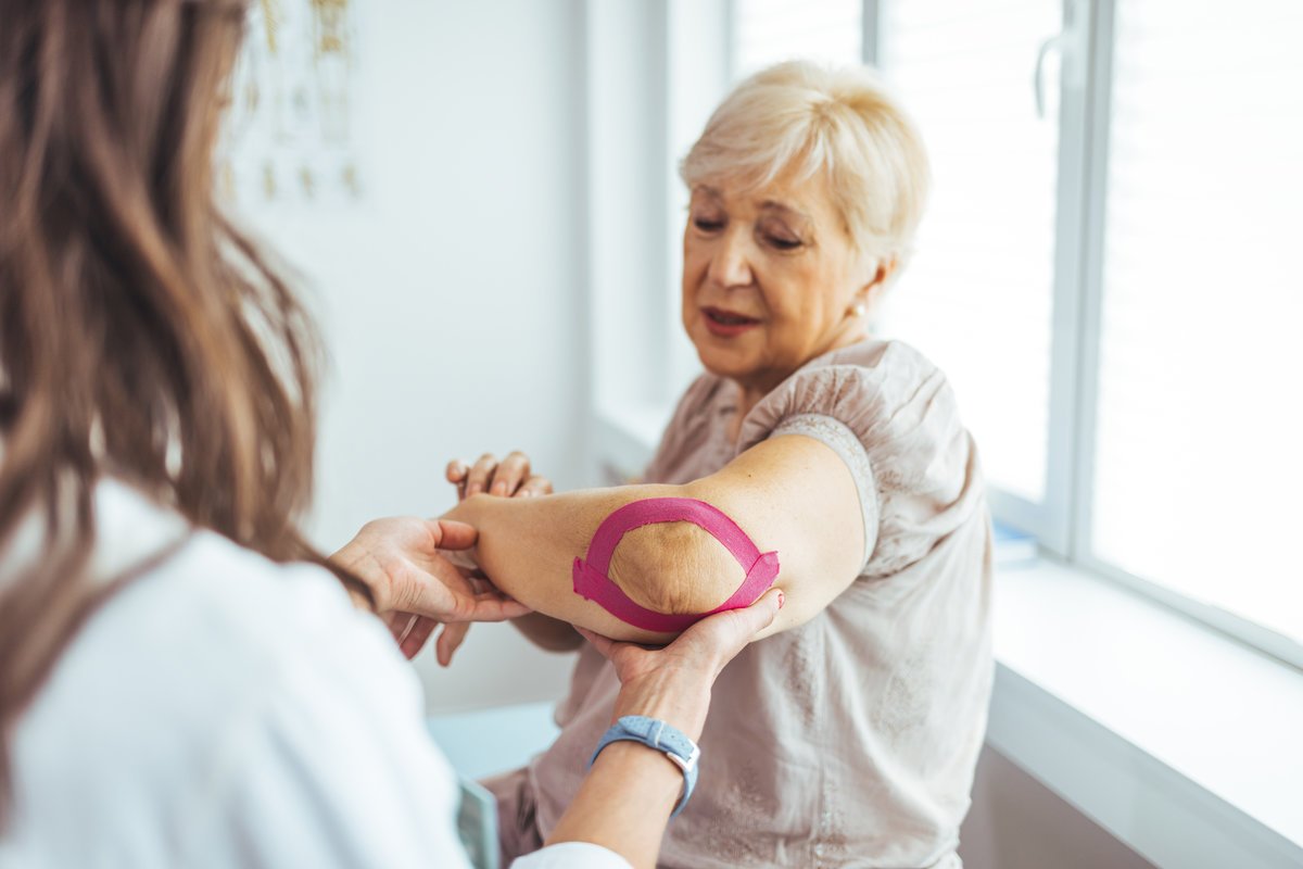 Kinesiology taping treatment with pink tape on senior patient injured arm. Woman hands apply kinesio treatment after muscle injury. A Modern rehabilitation physiotherapy worker with woman client