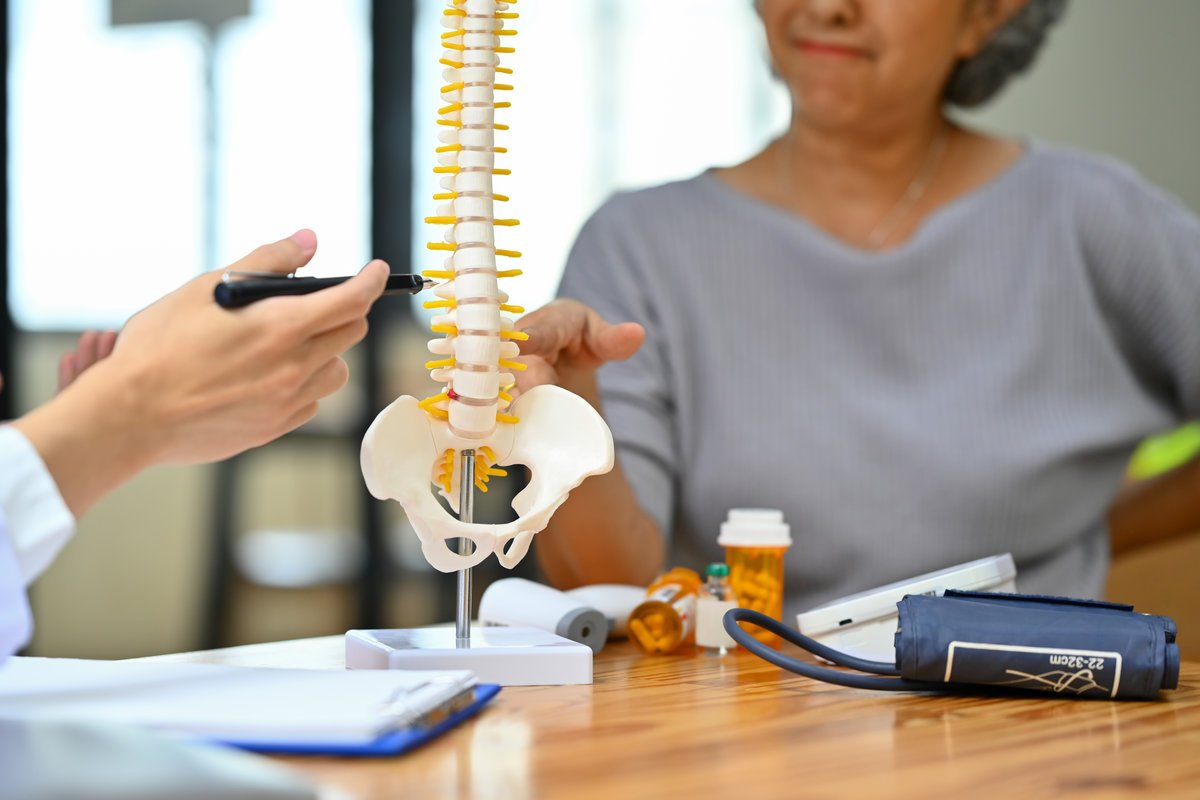 Orthopedist pointing at spine bone model, explaining the problem to mature female patient.